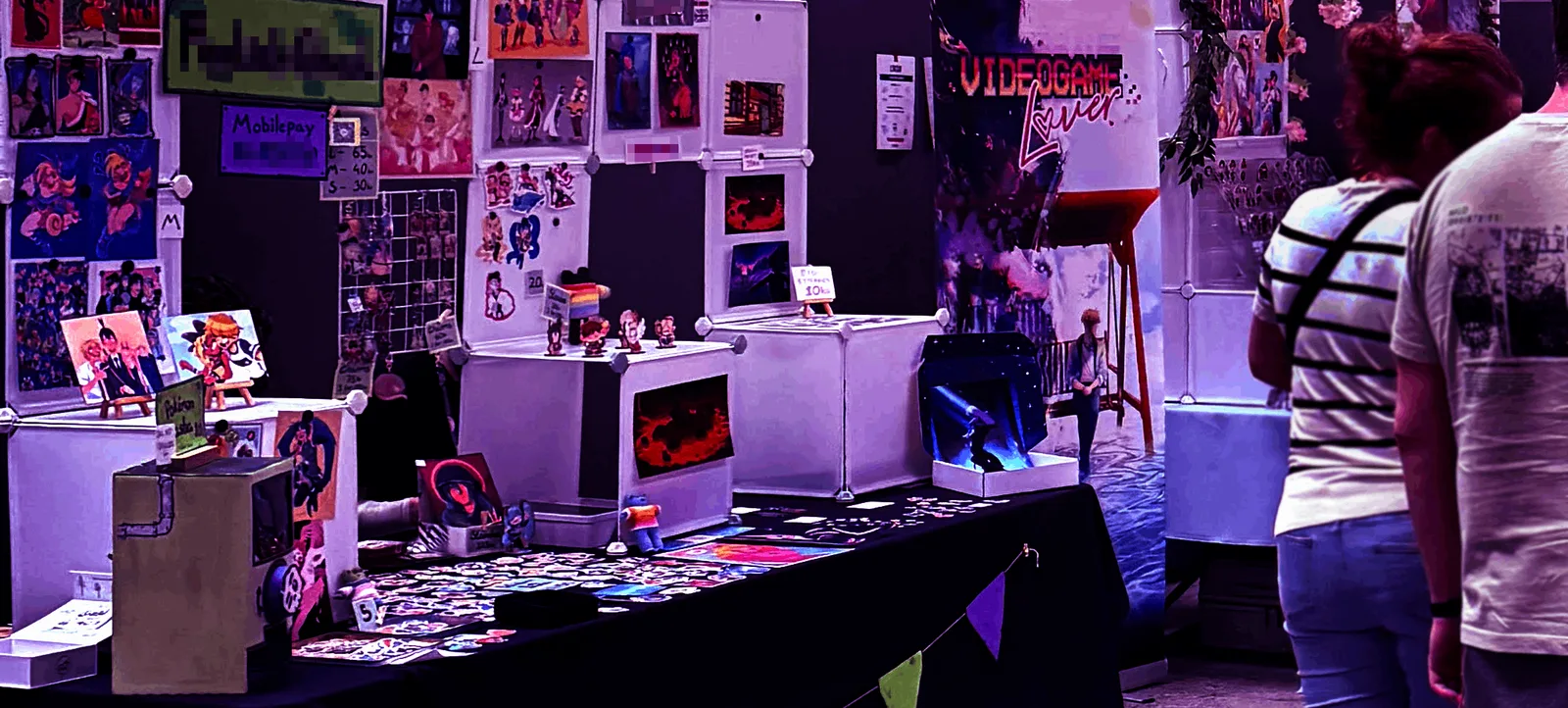 Stand i Artist Alley med prints, klistermærker og små figurer på væggen, mens to besøgende kigger forbi