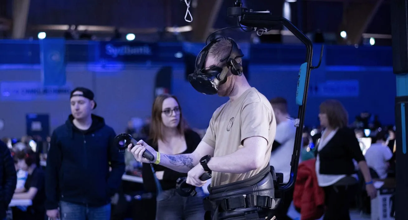 En gæst med VR-briller og controllere prøver et spil i en VR-rig på Expo, mens andre gæster ser til